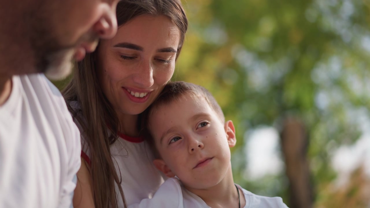 Primo piano di una donna e un bambino all'aperto, con il bambino che riposa sulla sua spalla. La scena mostra un'interazione familiare con luce naturale soffusa e vegetazione sfocata sullo sfondo. Entrambi indossano abiti bianchi