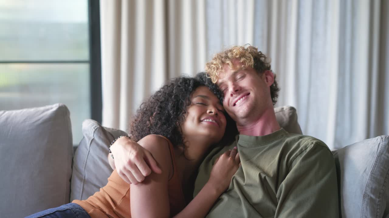 Diverse couple cuddling on sofa as woman leaning on man's chest, man wrapping arm for comfort