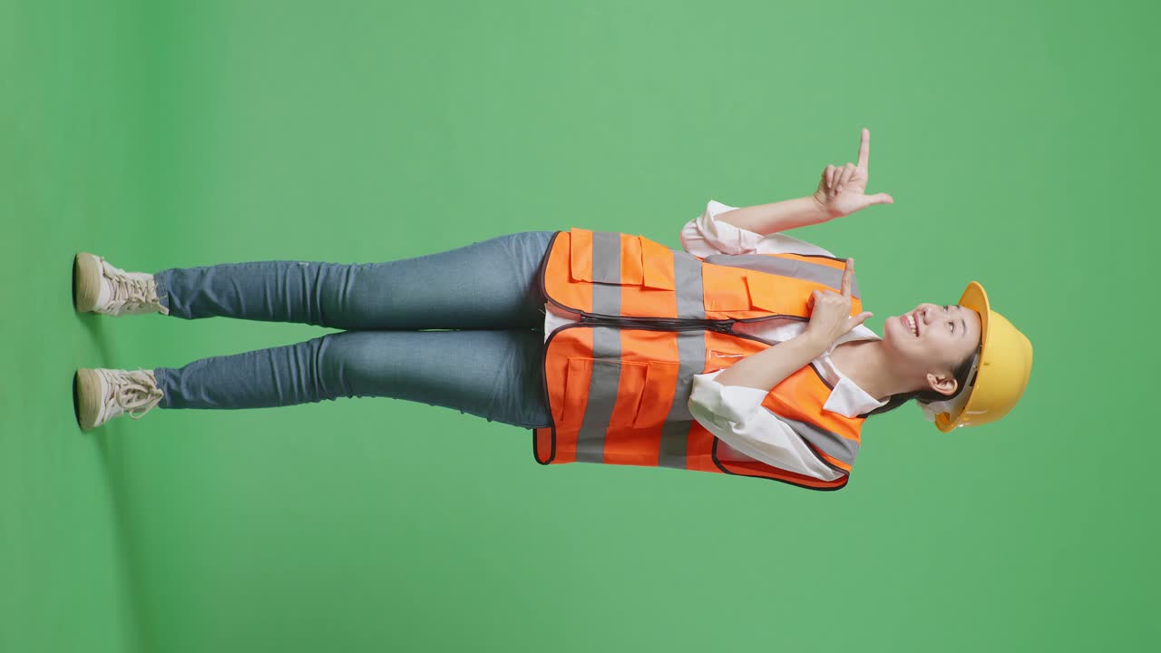 Full Body Of Asian Female Engineer With Safety Helmet Smiling And Pointing To Side While Standing In The Green Screen Background Studio