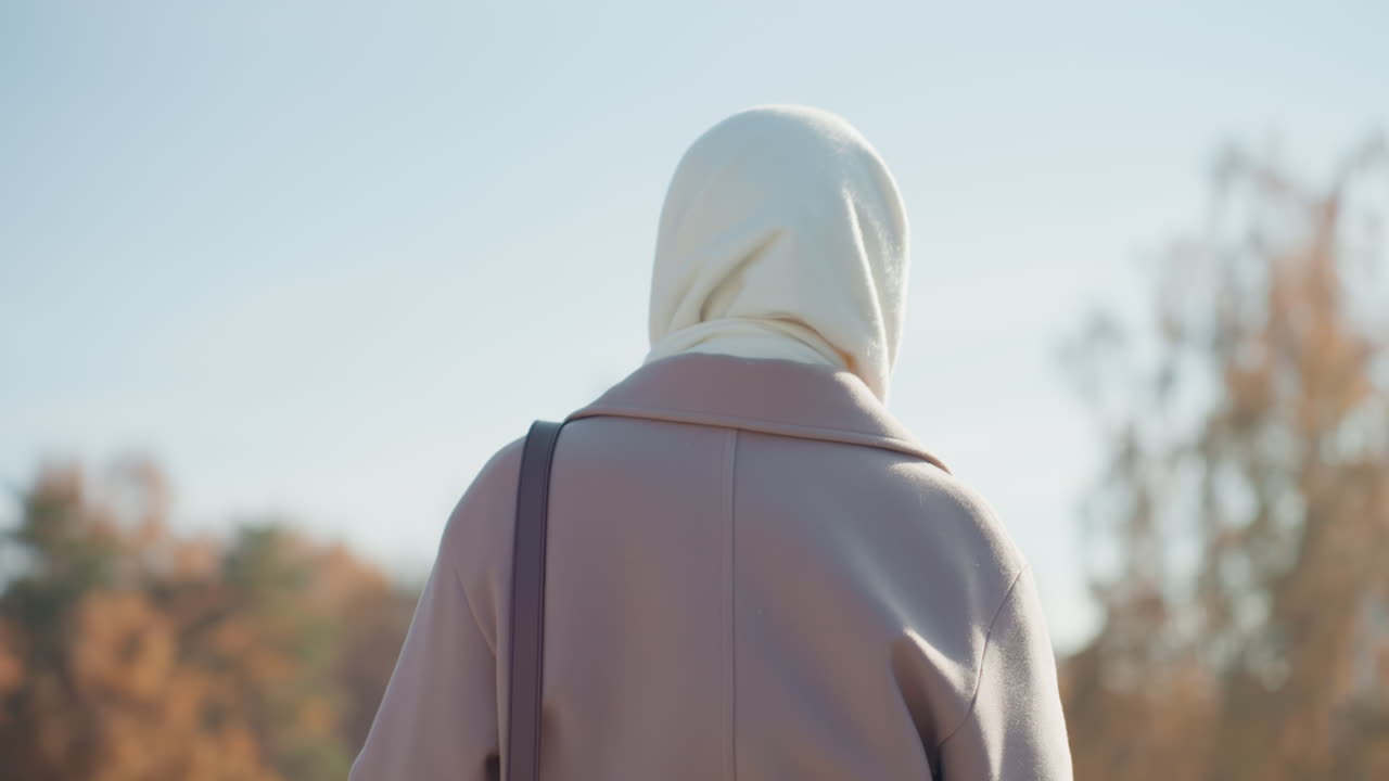 Back view woman wearing headscarf walking through city park lane, warm sunlight filtering through late fall trees, shoulder bag and candid urban mood, shallow depth and soft color grading