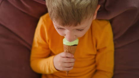close up aerial view of cute little boy sitting on pink bean bag relaxing indoors while holding mint chocolate ice cream cone and looking down at it with focus wearing yellow shirt