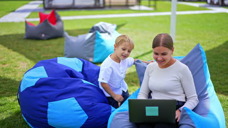little boy spending time outdoors in the green lawn. both look at laptop on lady's knees and talk. blurred backdrop.