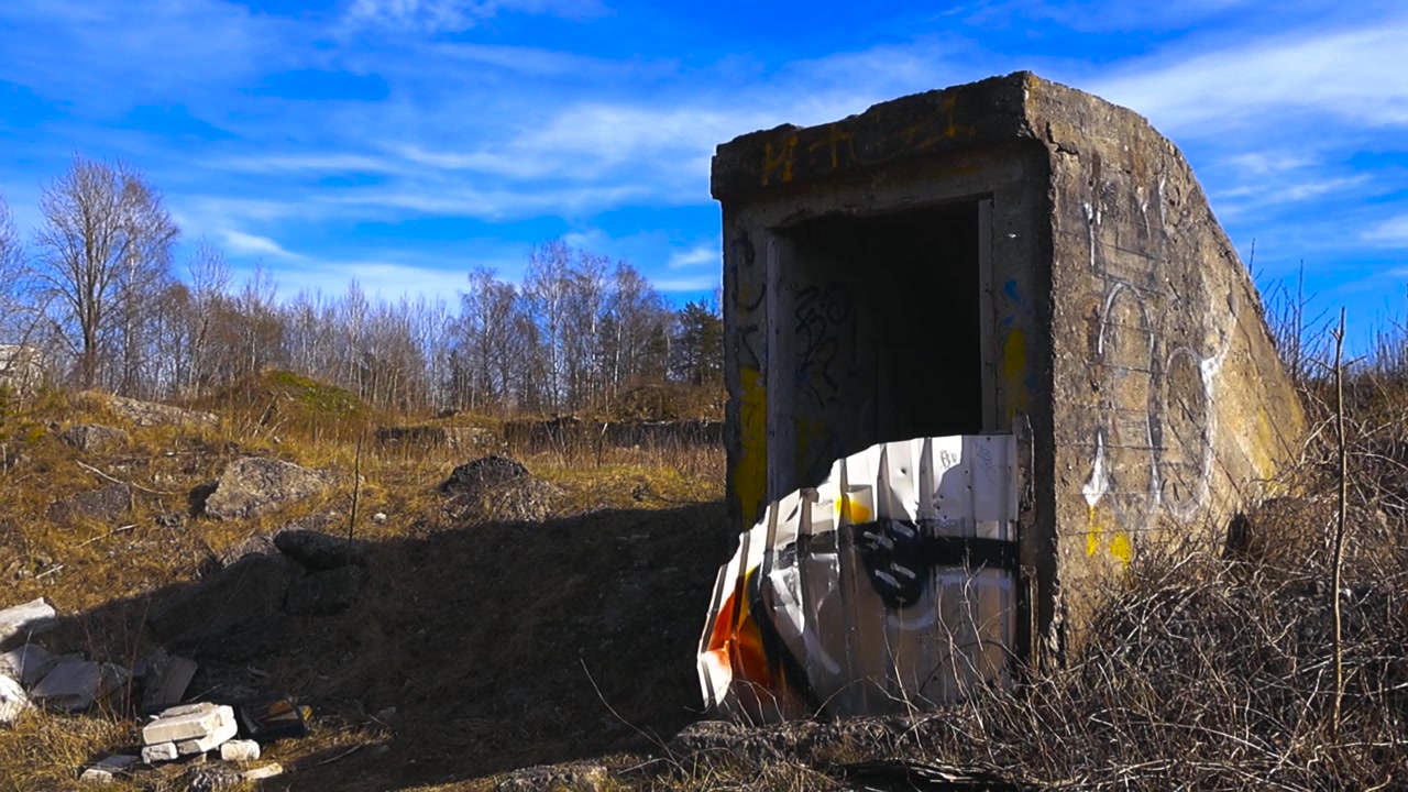 Antico ingresso di un bunker militare in cemento con graffiti sulle pareti, che si erge in un campo deserto sotto un cielo azzurro, a testimonianza di un'epoca passata e del passare del tempo.