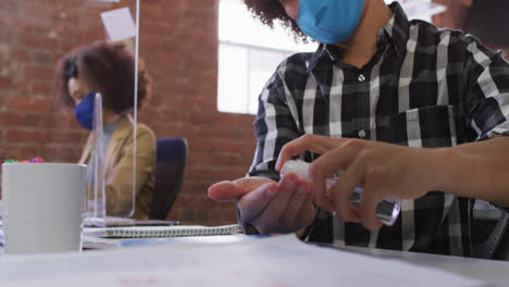 Mixed-race-businessman-disinfecting-hands-sitting-in-front-of-computer-wearing-face-mask