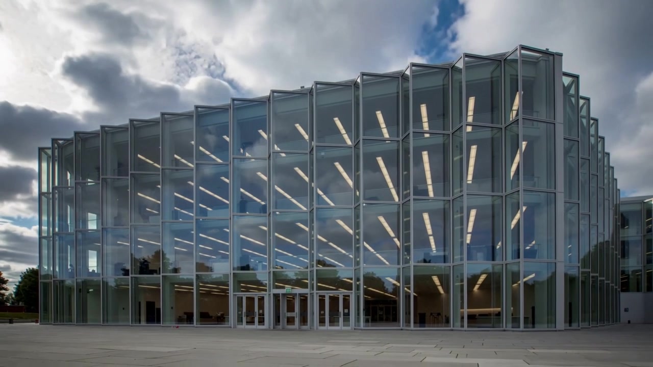 Nubes en movimiento proyectando reflejos sobre un edificio moderno con fachada de cristal en una plaza, con la capa de nubes cambiando.