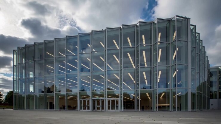 Nubes en movimiento proyectando reflejos sobre un edificio moderno con fachada de cristal en una plaza, con la capa de nubes cambiando.