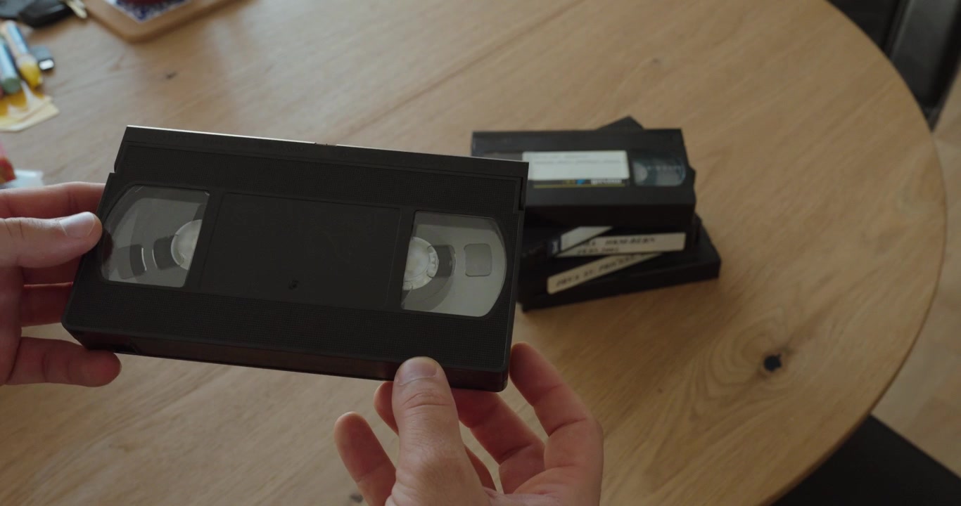 Close-up of hands holding and examining an old VHS cassette with more videotapes in the background. Ideal for vintage technology, analog video, nostalgia, home movies, archives, and media digitization
