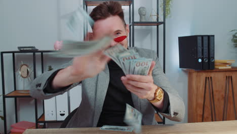 cheerful rich business man working on office laptop, wasting throwing money to camera, profit, win