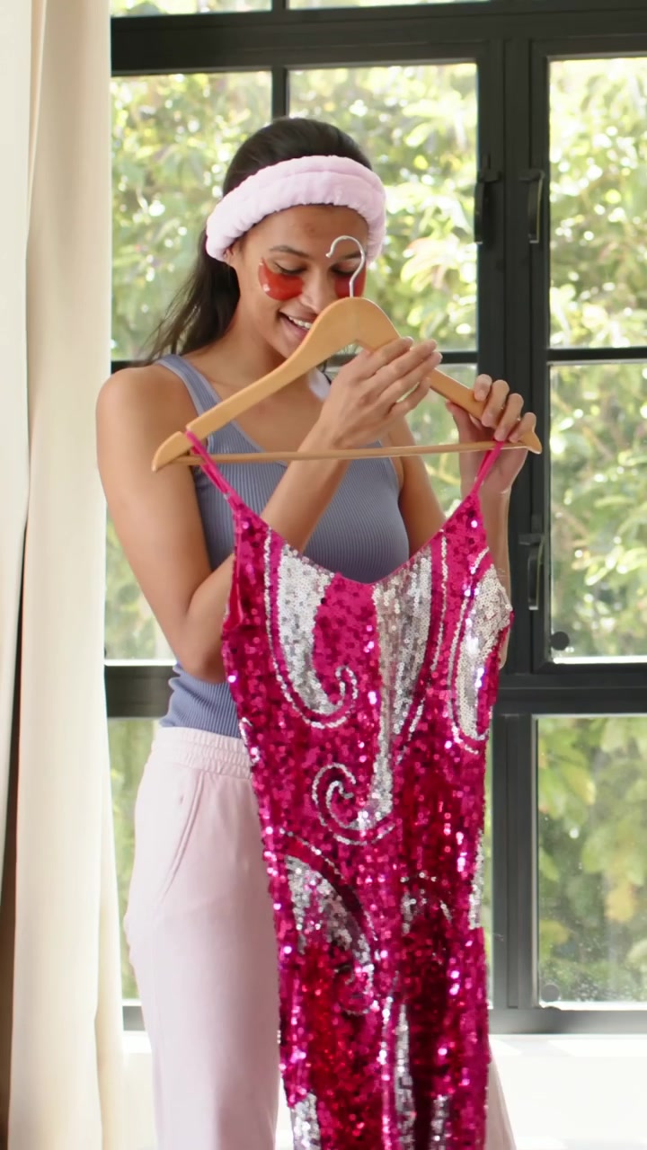 Video vertical: Sosteniendo un vestido de lentejuelas rosa plateado en una percha, mujer en camiseta de tirantes revisando el vestido junto a la ventana.