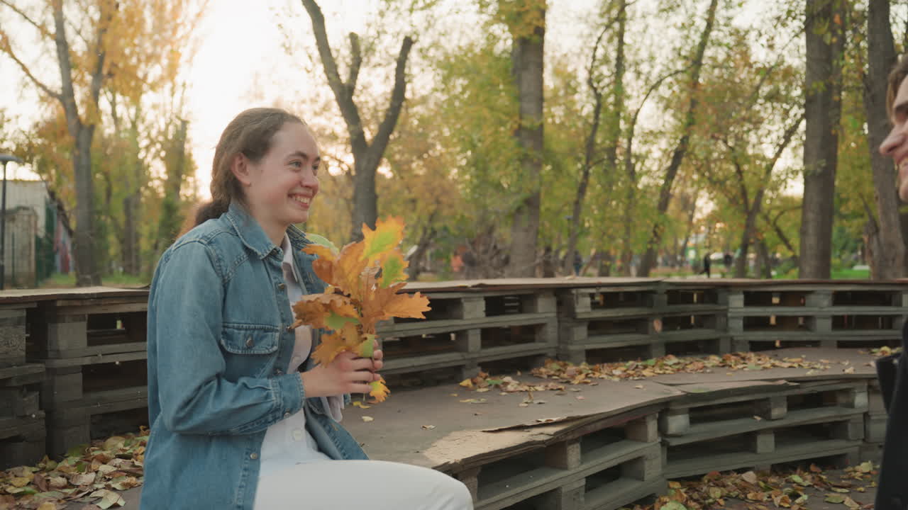 Donna bianca che riceve un bouquet di foglie autunnali su una panchina del parco, reazione in primo piano con lentiggini e giacca di jeans, luce calda e dorata, il partner che offre le foglie con un gesto giocoso da appuntamento, con un sorriso tenero e grato.