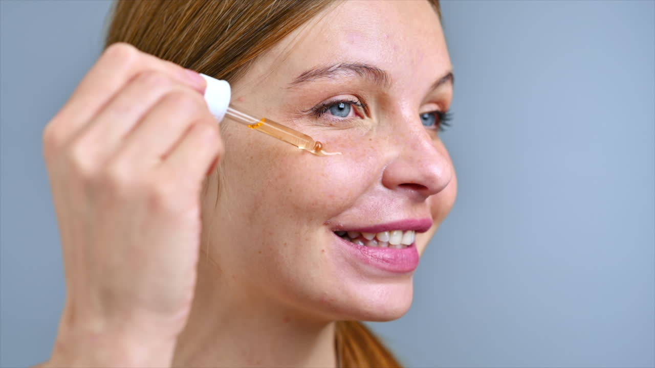 A young caucasian smiling woman is applying a cosmetic oil to her face. Slow motion