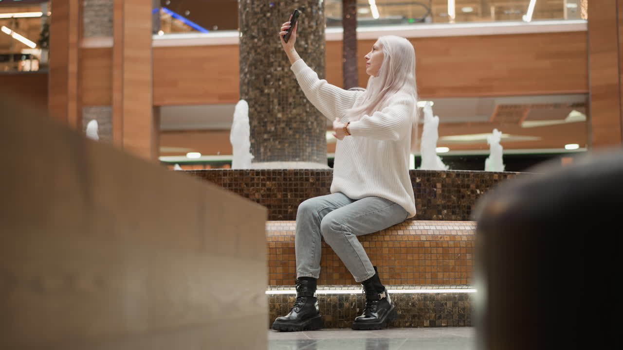 Chic mom adjusting long hair sits on tiled bench beside indoor mall fountain raising mobile phone to pose capture fountain jets and ambient lighting while shoppers stroll past glossy retail windows