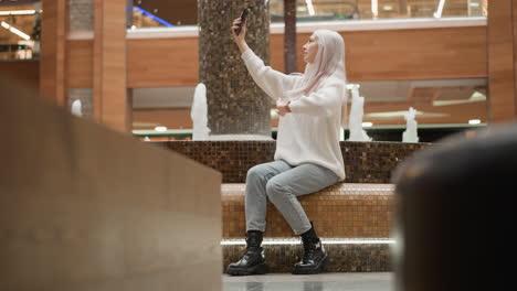 chic mom adjusting long hair sits on tiled bench beside indoor mall fountain raising mobile phone to pose capture fountain jets and ambient lighting while shoppers stroll past glossy retail windows