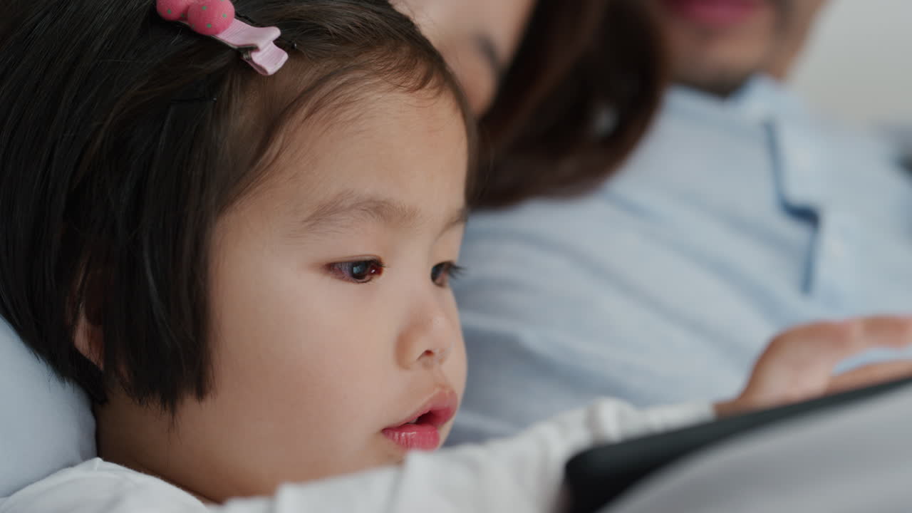 가족과 함께 태블릿 컴퓨터를 사용하는 귀여운 작은 아시아 소녀는 터치 스크린 장치에서 게임을 즐기고 집에서 함께 휴식을 취합니다 4k