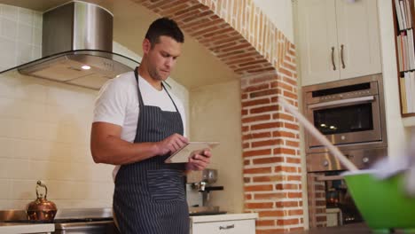 Kaukasischer-Mann-Trägt-Schürze,-Während-Er-Zu-Hause-In-Der-Küche-Ein-Digitales-Tablet-Benutzt