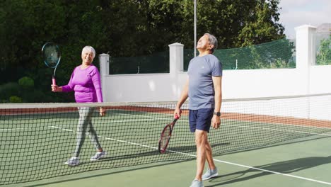 video of happy biracial senior couple walking with rackets on tennis court