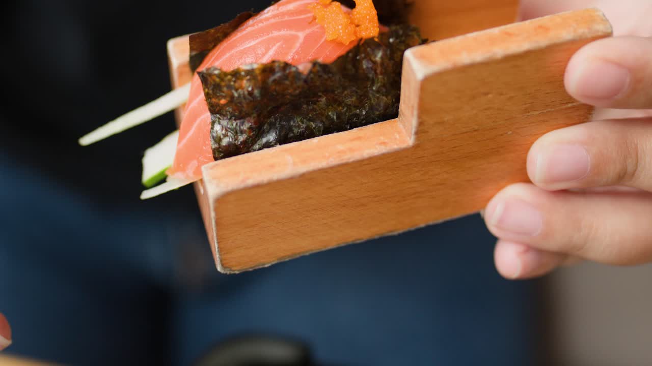 Primo piano di una persona che solleva un involtino di sushi al salmone fresco da un supporto di legno con illuminazione naturale soffusa