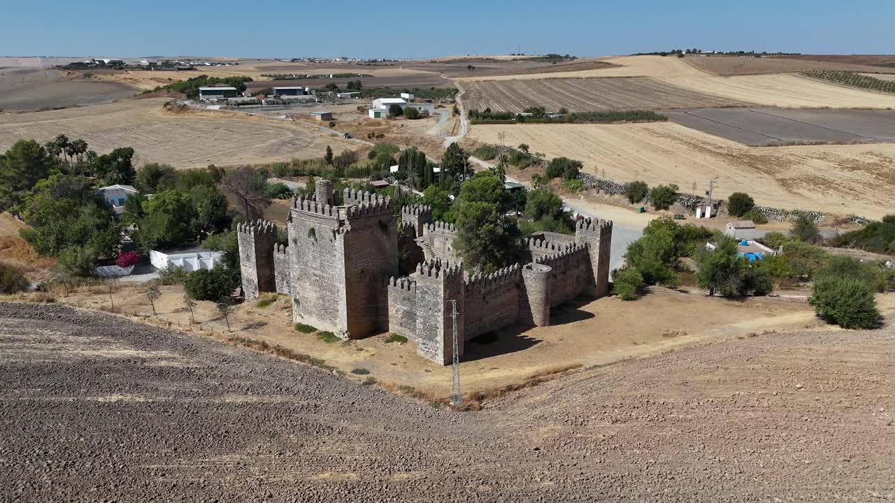 Drone tracking back and rising over Castillo de las Aguzaderas near El Coronil, Seville, Spain, gradually revealing its stone walls, towers, and the expansive, sunlit, dry Andalusian landscape