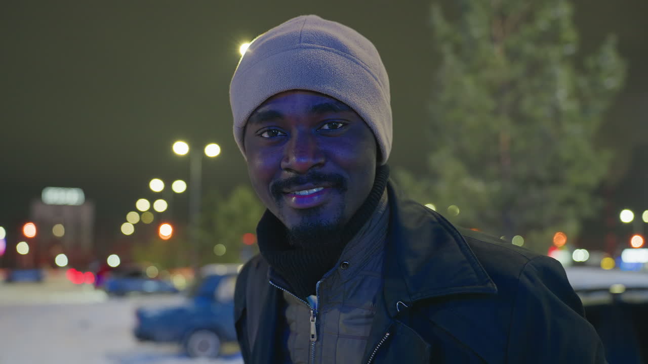 Vista laterale di un uomo con cappello invernale all'aperto di notte che espira aria fredda visibile, si gira con un dolce sorriso sul volto con luci della città sfocate e sfondo innevato