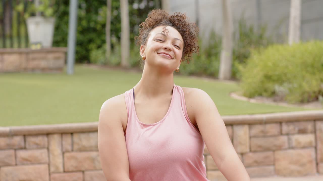 Vendo a câmera se aproximar, mulher de regata rosa se alongando, rindo em um tapete de yoga roxo no quintal
