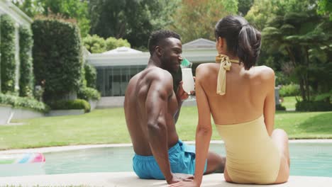 happy diverse couple wearing swimming suits and holding hands at swimming pool in garden
