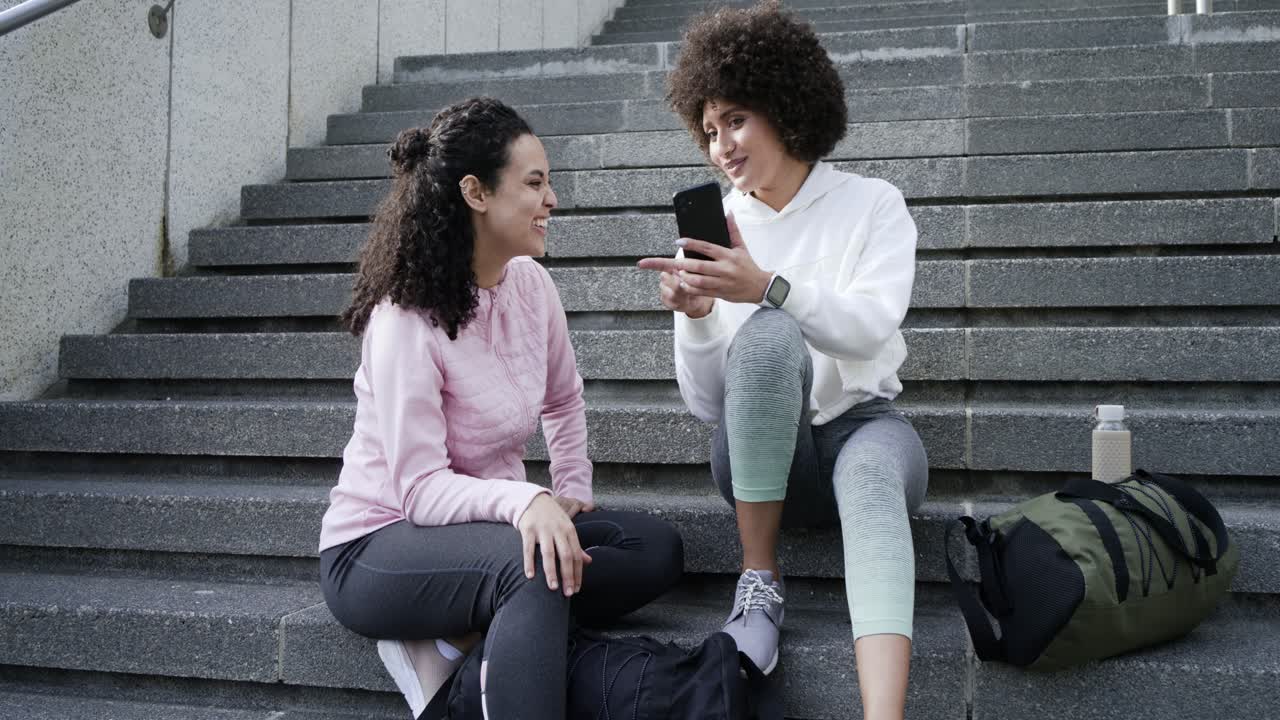 Amiche di varie etnie che mostrano uno smartphone, chiacchierando animatamente, scorrendo e annuendo sui gradini di cemento.