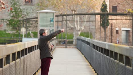 A-man-stands-on-a-bridge-with-a-metal-fence-and-points-off-into-the-distance