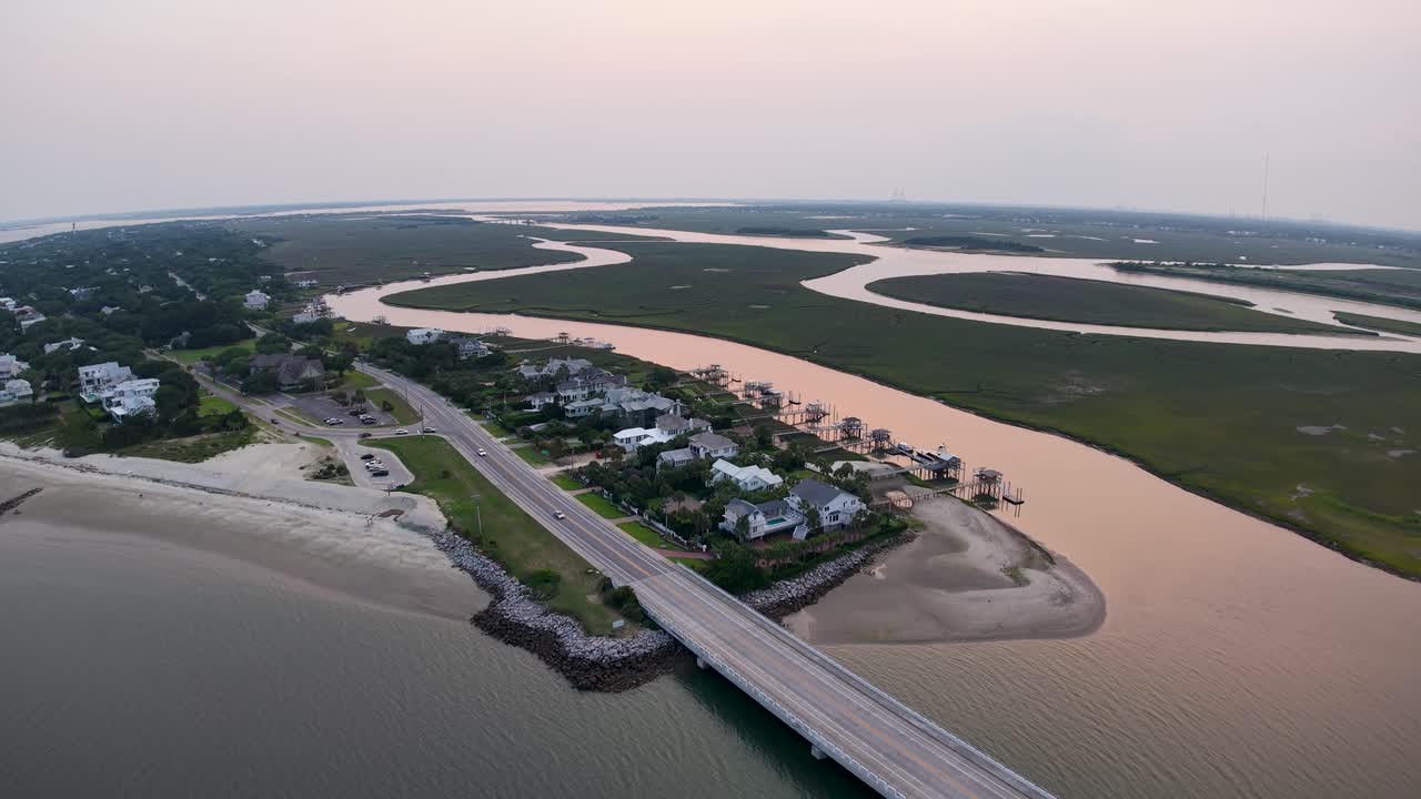Drone footage of a long bridge crossing over a tidal river to a small coastal community in Charleston, South Carolina, showing houses along the shoreline, wetlands and roads during sunset