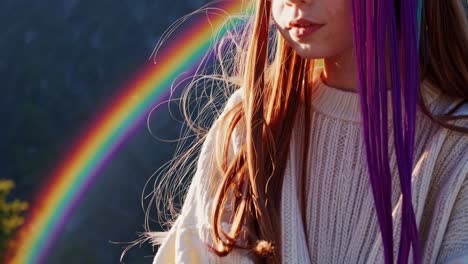 redheaded woman wearing vibrant rainbow hair extensions, gazing downward against blurred rainbow backdrop, representing inclusivity and personal expression
