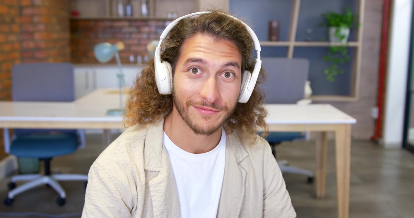 Homem na casa dos 20 anos com fones de ouvido em uma mesa no escritório, olhando para cima depois de ler e sorrindo para a câmera.