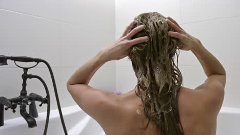 una mujer desnuda sentada cómoda en la bañera mientras aplica productos para el cabello en su largo cabello con las manos