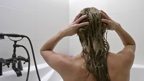 a naked woman sitting comfortable in the bathtub while applying hair product in her long hair using her hands