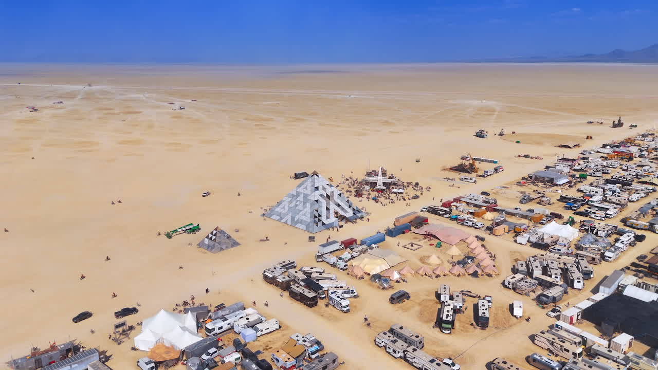 Large geometric pyramids on open playa. Metallic pyramid structures rise beside camps and bikes across the vast desert floor