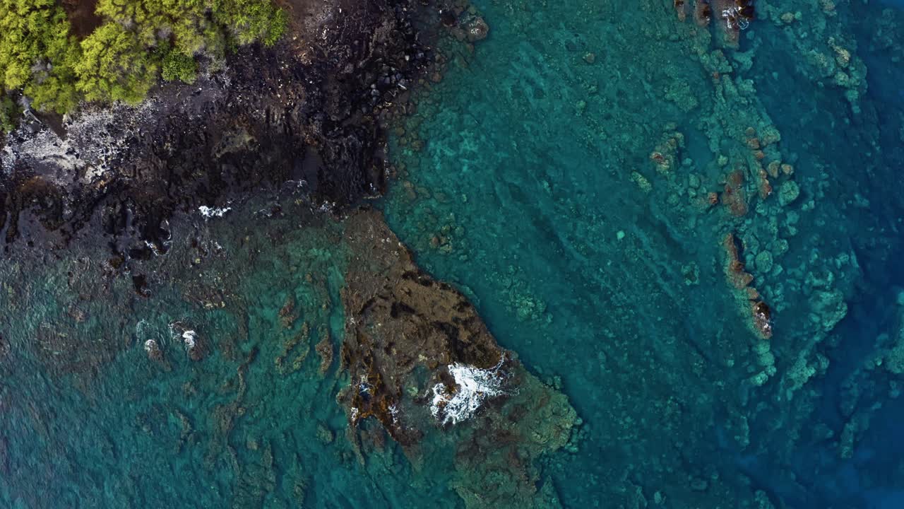 Rugged coastal rocks meet transparent turquoise waters where coral reefs shimmer beneath the surface, revealing textures and patterns shaped by ocean currents