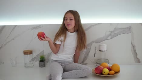 little girl with long fair hair eating red apple in bright, modern kitchen while sitting on a counter. bowl of fresh fruits next to her. girl enjoying fresh apple