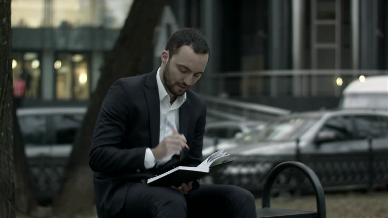 Young bearded business man thinking and wrighting notes at green park, traffic behind