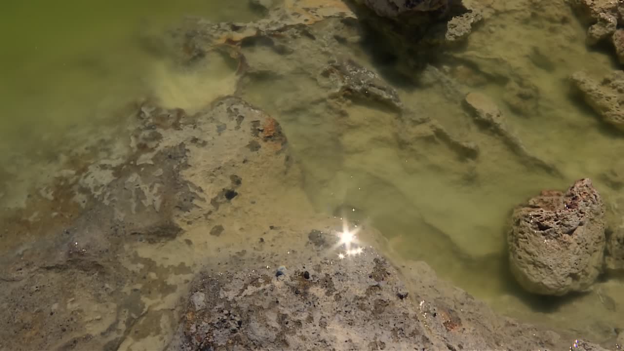 Rocky lakebed in shallow green water with a bright sun reflection near the surface at Mono Lake, California