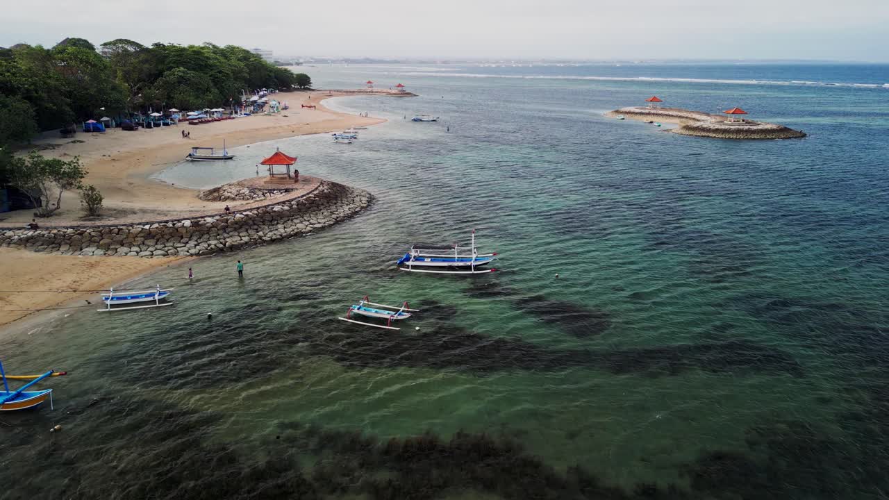 Aerial footage captures Sanur Beach Bali with turquoise ocean, sandy coastline, protective coastal barriers, and a colorful traditional fishing boat highlighting local culture and scenery