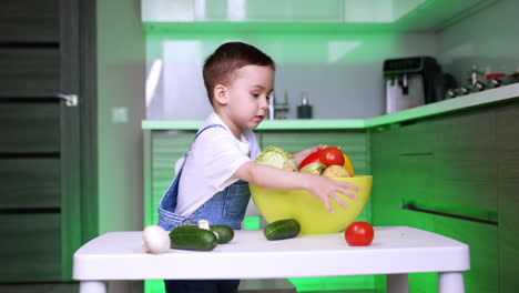 baby wearing jeans romper puts vegetables into a bowl. child learns the veggies and healthy eating habits.