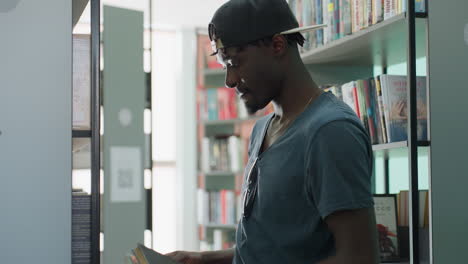 hombre con gorra informal y camiseta sujetando dos libros en el pasillo de una biblioteca, devolviendo un libro de tapa roja a la estantería mientras se queda con otro, rodeado de filas de libros coloridos bajo una luz suave, concentrado en la tarea
