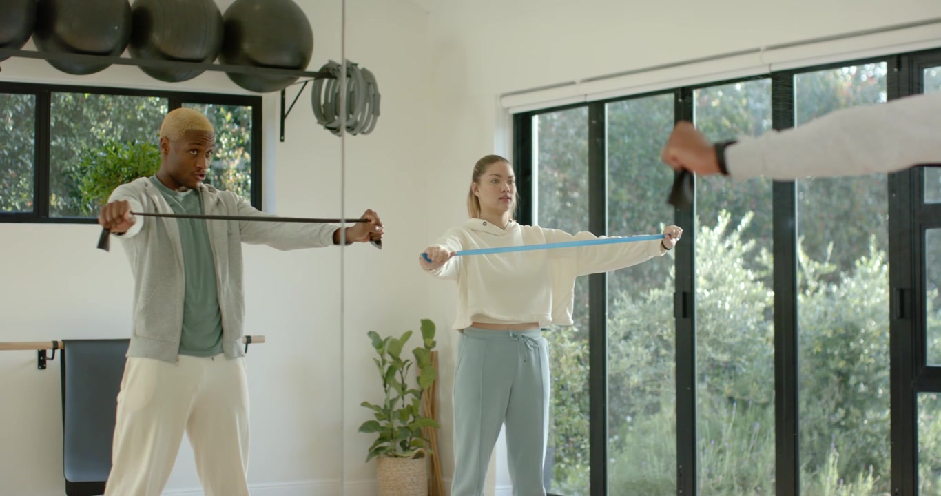 Diverse workout partners performing band pulls in sync at home gym, man glancing to lead timing
