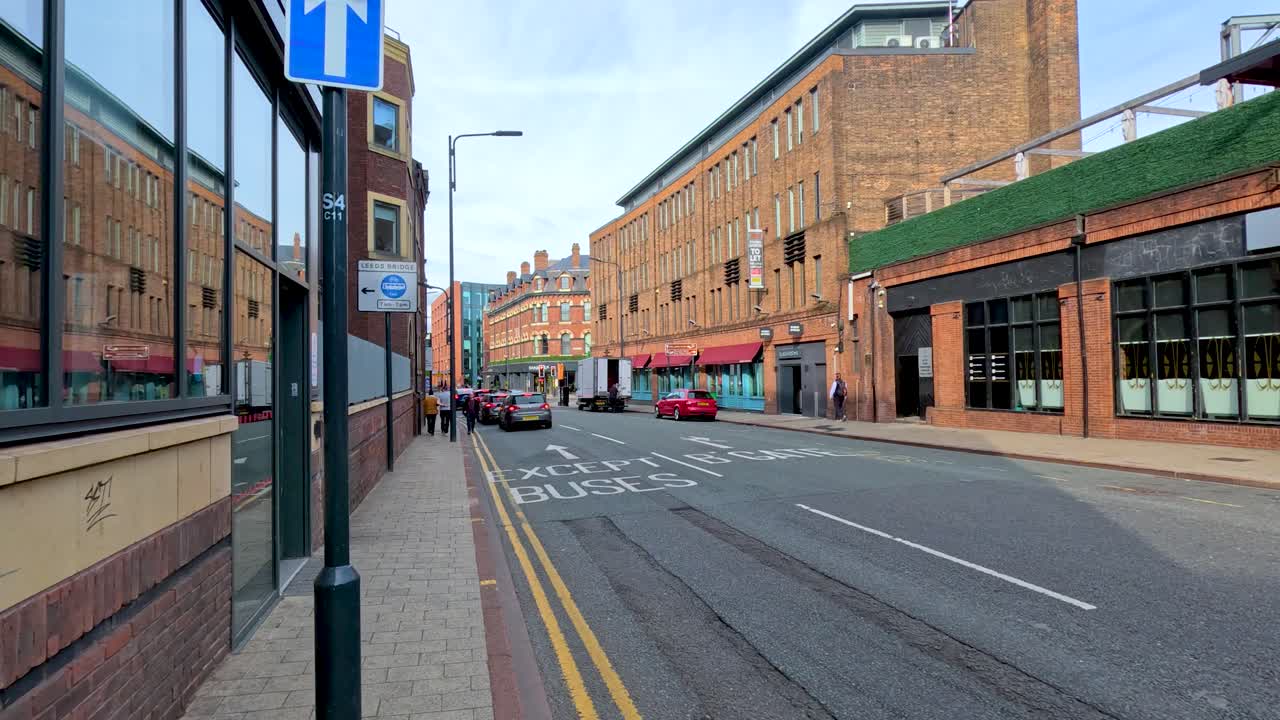 Camera moves steadily down a quiet urban street beside a large brick building in Leeds, England, under natural daylight with clear visibility