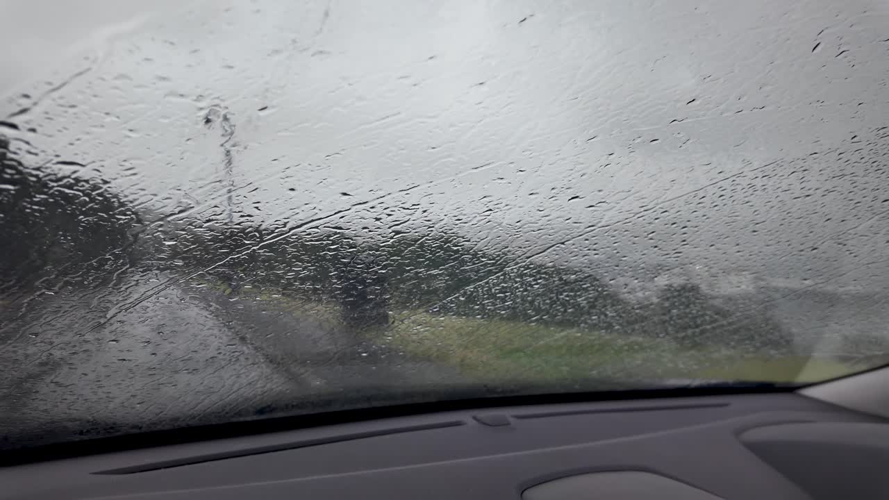 Rain-soaked windshield blurs the countryside, raindrops streak the glass, captured from inside a car during a storm