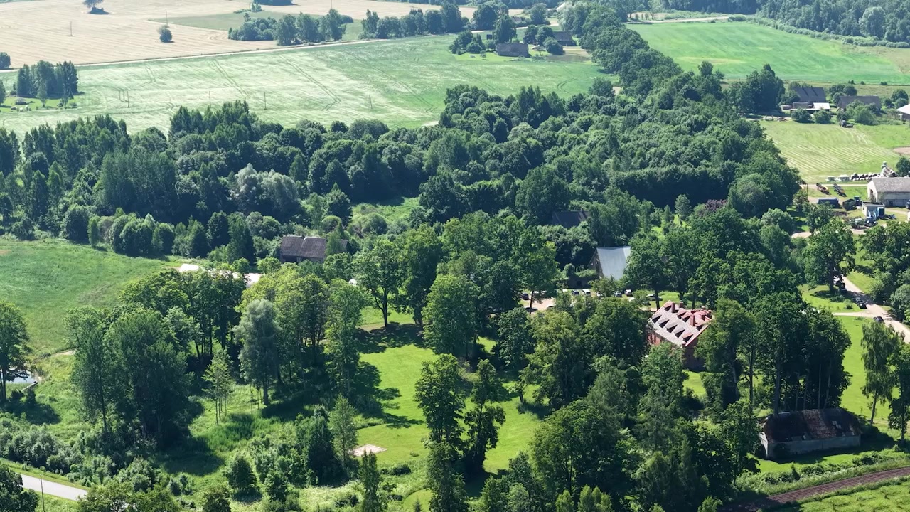 Dronekuva vihreistä maaseudun pelloista, hiekkatiestä, puuriveistä, metsälaikuista, heinäpelloista ja hajallaan olevista maatalousrakennuksista aurinkoisessa maaseutumaisemassa.
