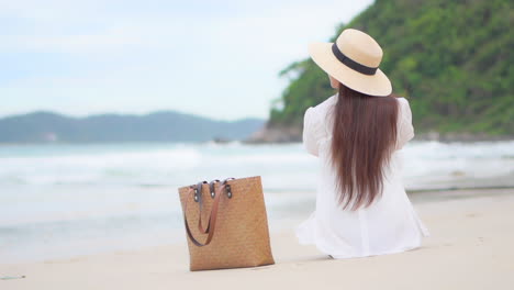 Mujer-Irreconocible-Sentada-En-Una-Playa-De-Arena-Blanca-Frente-Al-Mar-Con-Un-Pantalones-De-Blusa-Blanca-Y-Un-Sombrero-De-Paja,-Bolsa-De-Playa,-Naturaleza-Tropical-De-La-Isla-Hawaii-En-El-Fondo,-Vista-Trasera-En-Cámara-Lenta