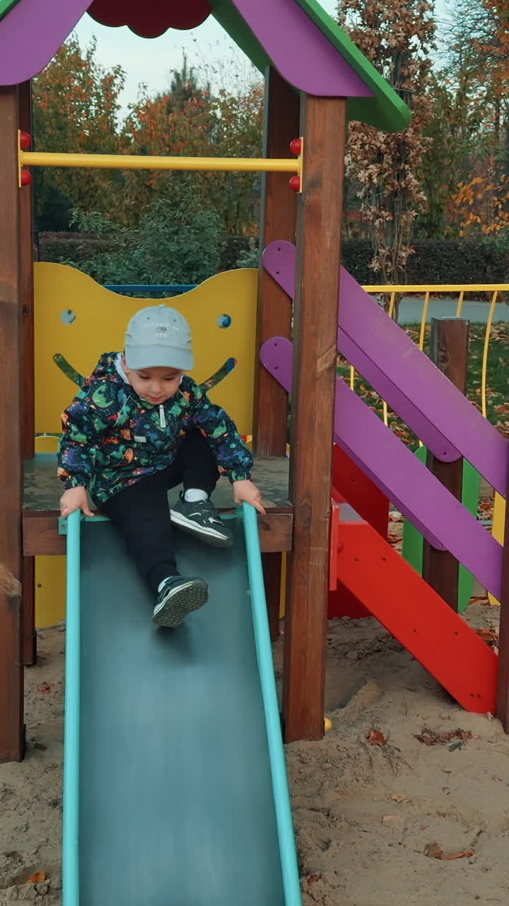 Boy wearing jacket and cap waving on the horizontal bar. Kid sits down on the slide and taps his feet happily. Playground backdrop. Vertical video