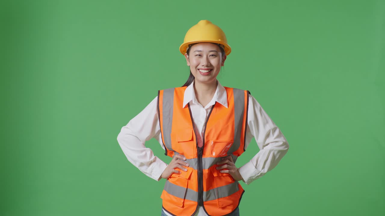 Asian Female Engineer Wearing Safety Helmet Smiling To Camera While Standing With Arms Akimbo In The Green Screen Background Studio