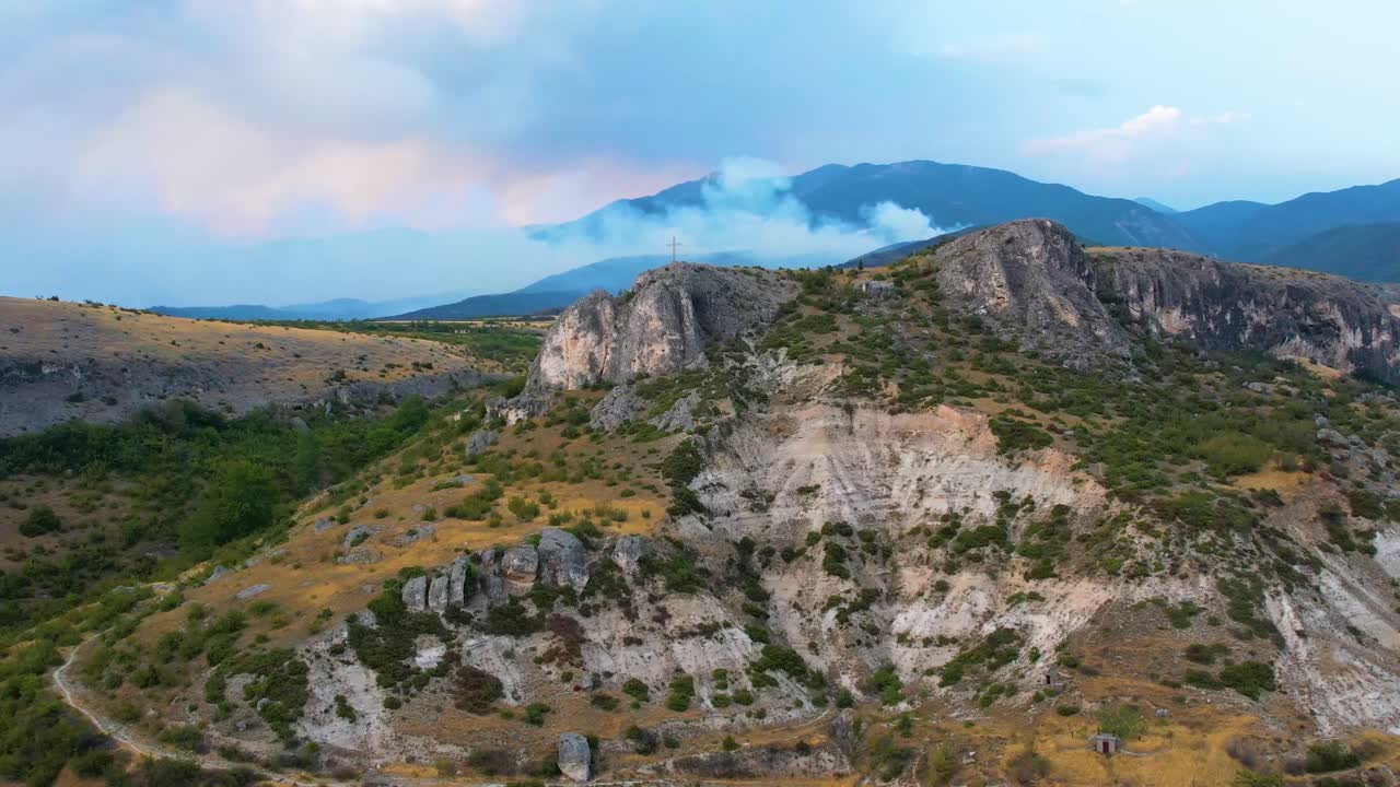 Camera reverse begins distant from monument, capturing vineyards, dry hills and valley as grey wildfire smoke from burning Pirin Mountain forests drifts across skyline during breaking summer heat