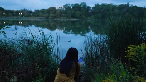 Mujer-Joven,-Sentada-Junto-Al-Lago-Al-Amanecer,-Al-Teléfono,-Pato-Nadando-Cerca