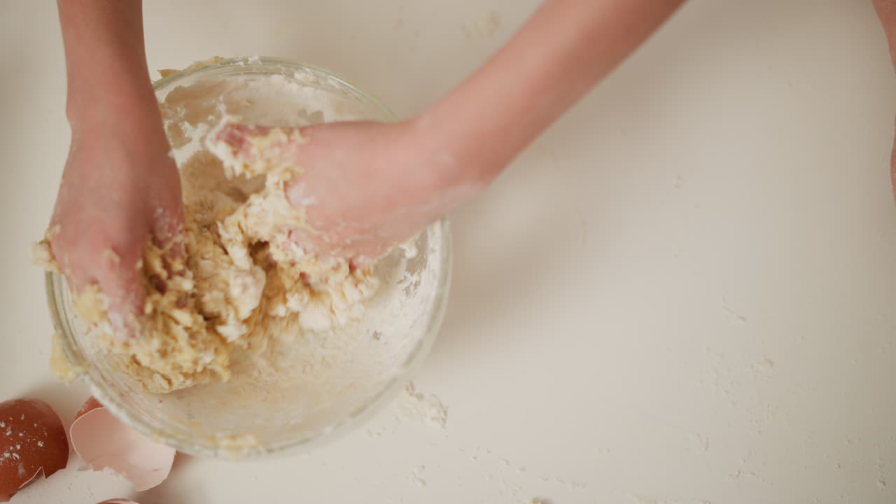 Overhead view of fair-skinned hands kneading soft dough in glass bowl on floured surface with broken brown eggshells nearby, fingers coated with flour during messy and engaging baking